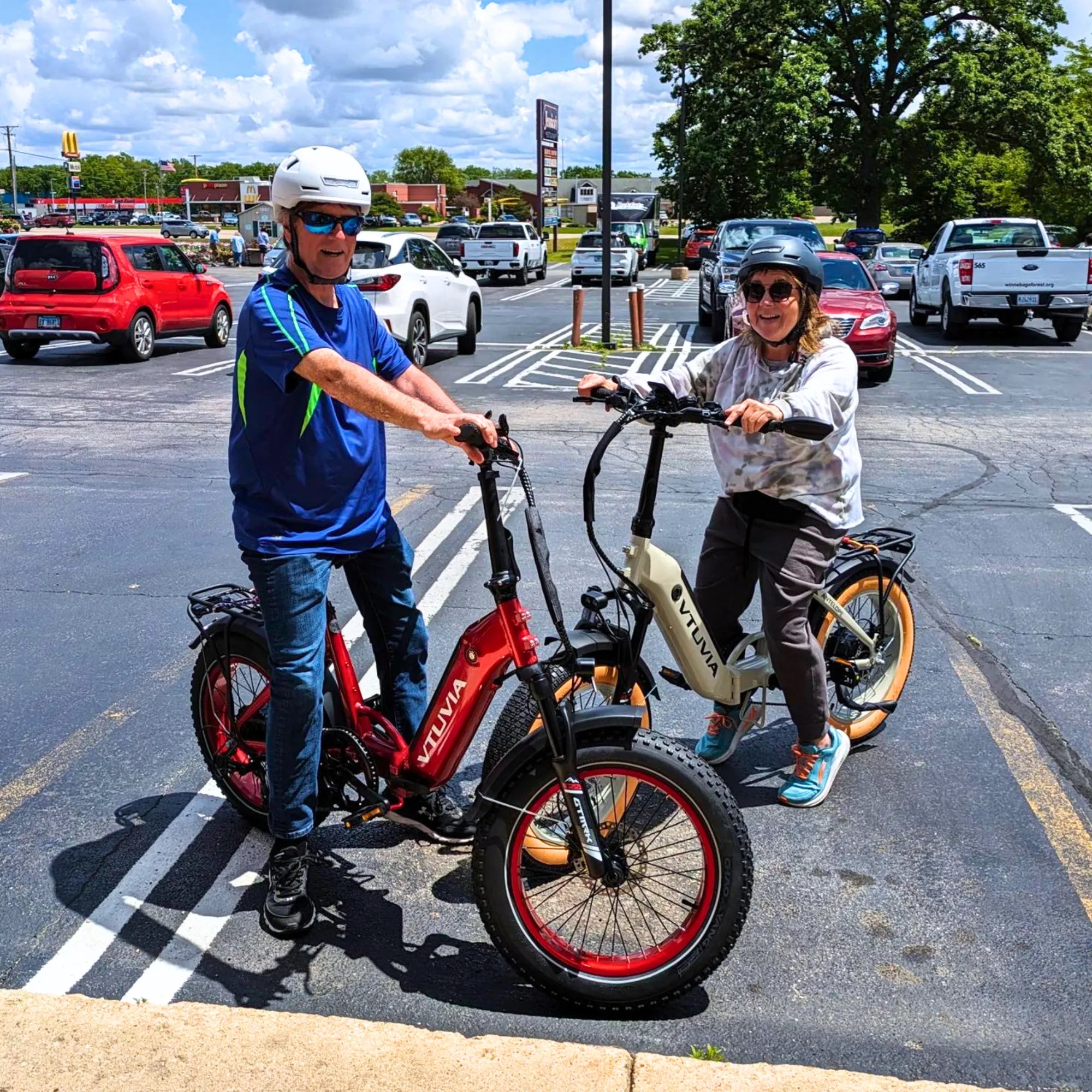 Two smiling seniors riding red VTUVIA SX20 and white Antelope low step-through fat tire electric bikes in a city parking lot, focusing on comfort and ease of use.
