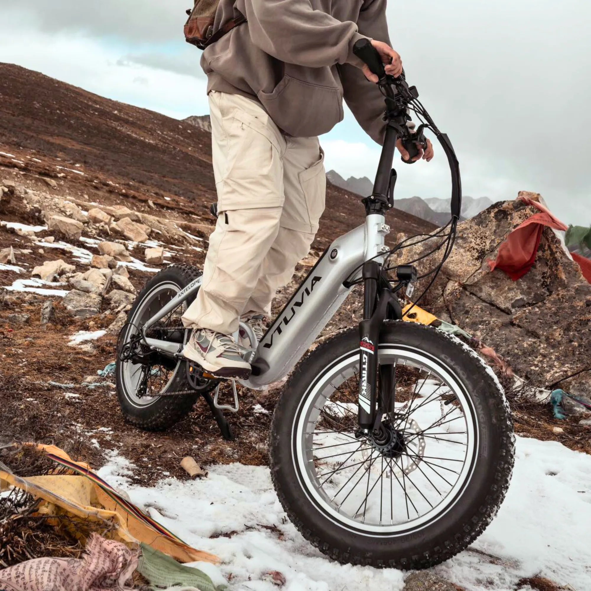 Rider on a silver VTUVIA SX20 fat tire electric bike conquering snow covered and rocky mountain terrain, highlighting its 1200W peak power.