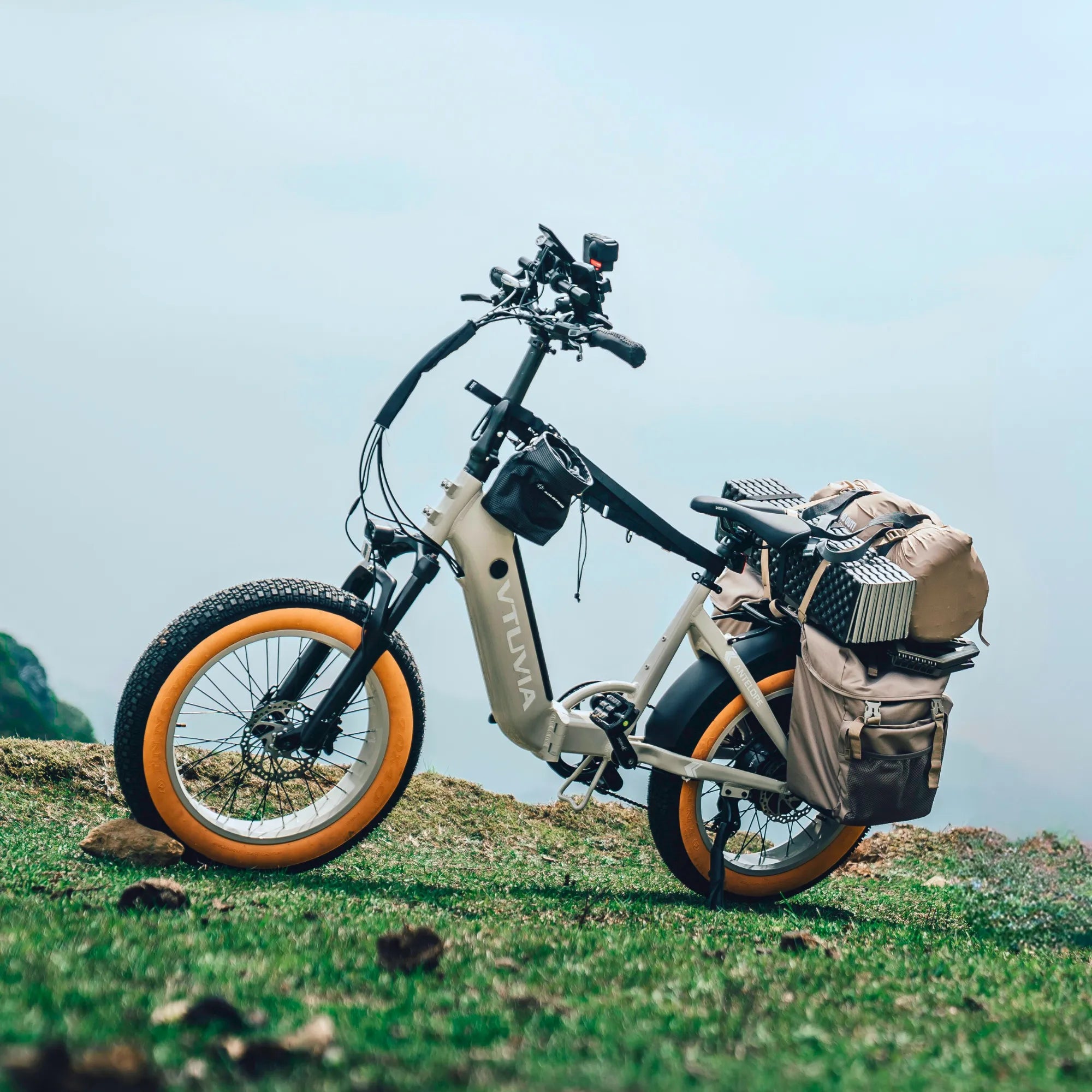 VTUVIA Antelope sand colored electric bike with yellow tires fully loaded with camping gear and panniers on a grassy hill, showcasing high payload capacity.